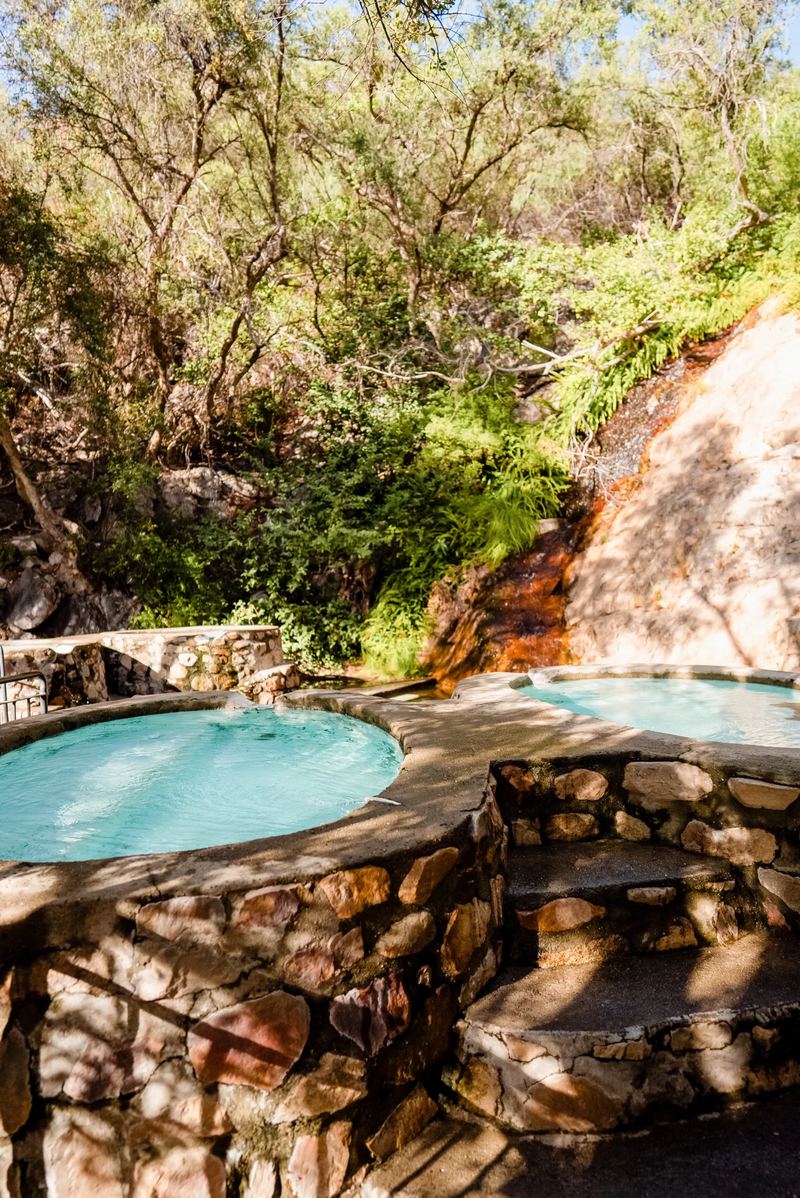 A small round stone hot pool beside an orange rock face in the green kloof at The Baths.