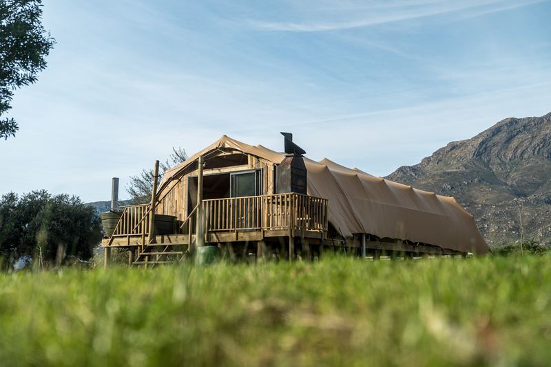 A Wolfkop glamping villa on its raised wooden deck, canvas roof and stone chimney, mountains behind.